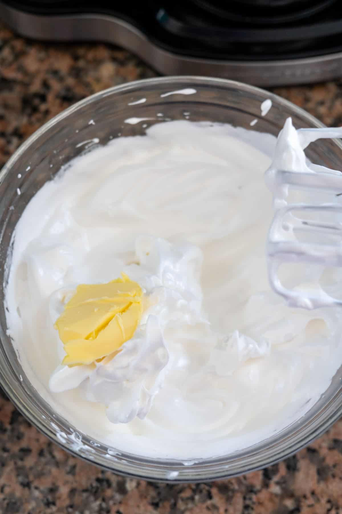 A glass bowl filled with whipped cream or thick white mixture, reminiscent of the smooth frosting atop lemon lavender cupcakes, is topped with a small dollop of yellow butter. A metal whisk is partially visible on the right side.