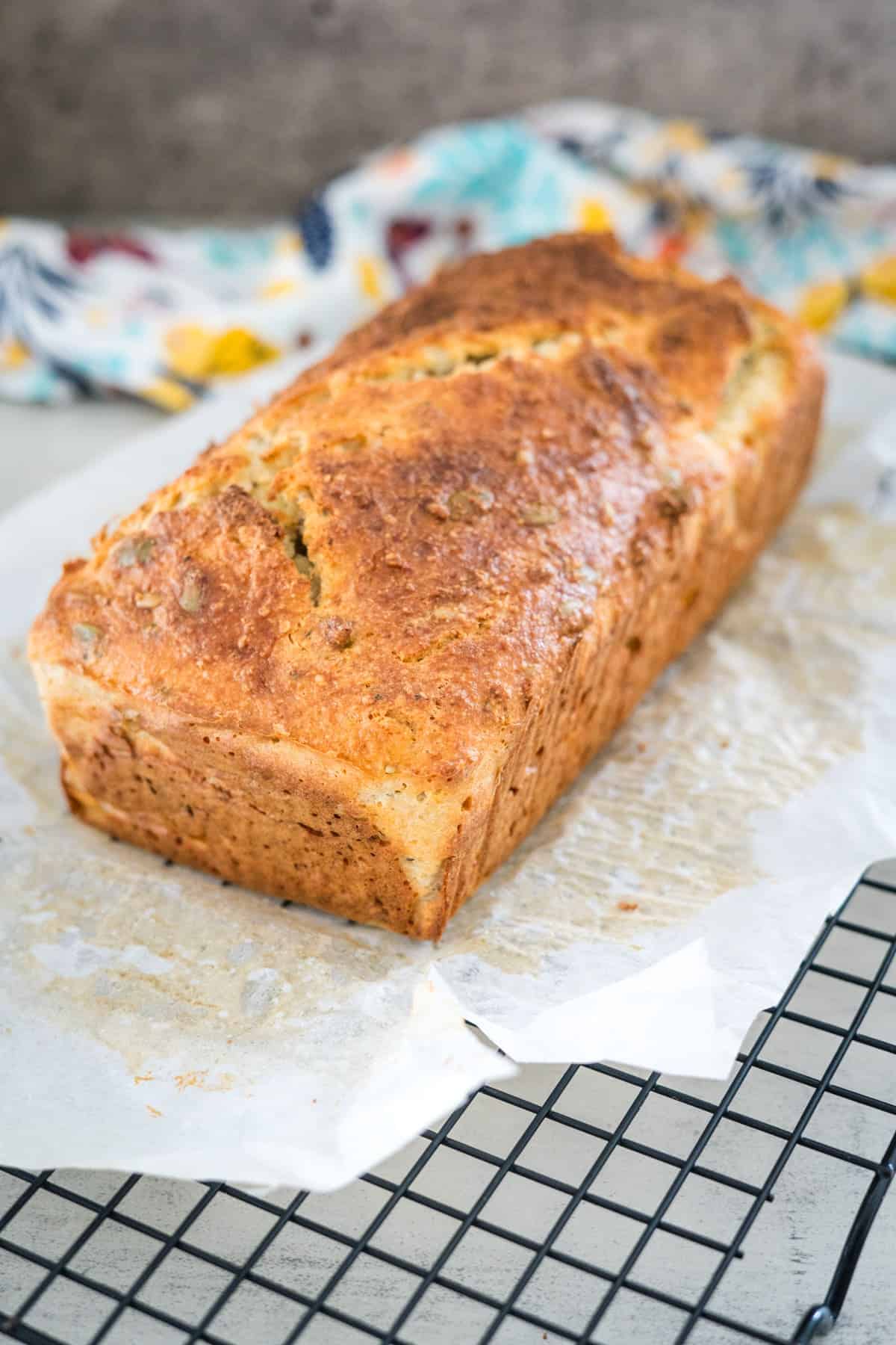 A loaf of freshly baked banana bread rests on parchment paper atop a cooling rack, with a floral-patterned cloth in the background.