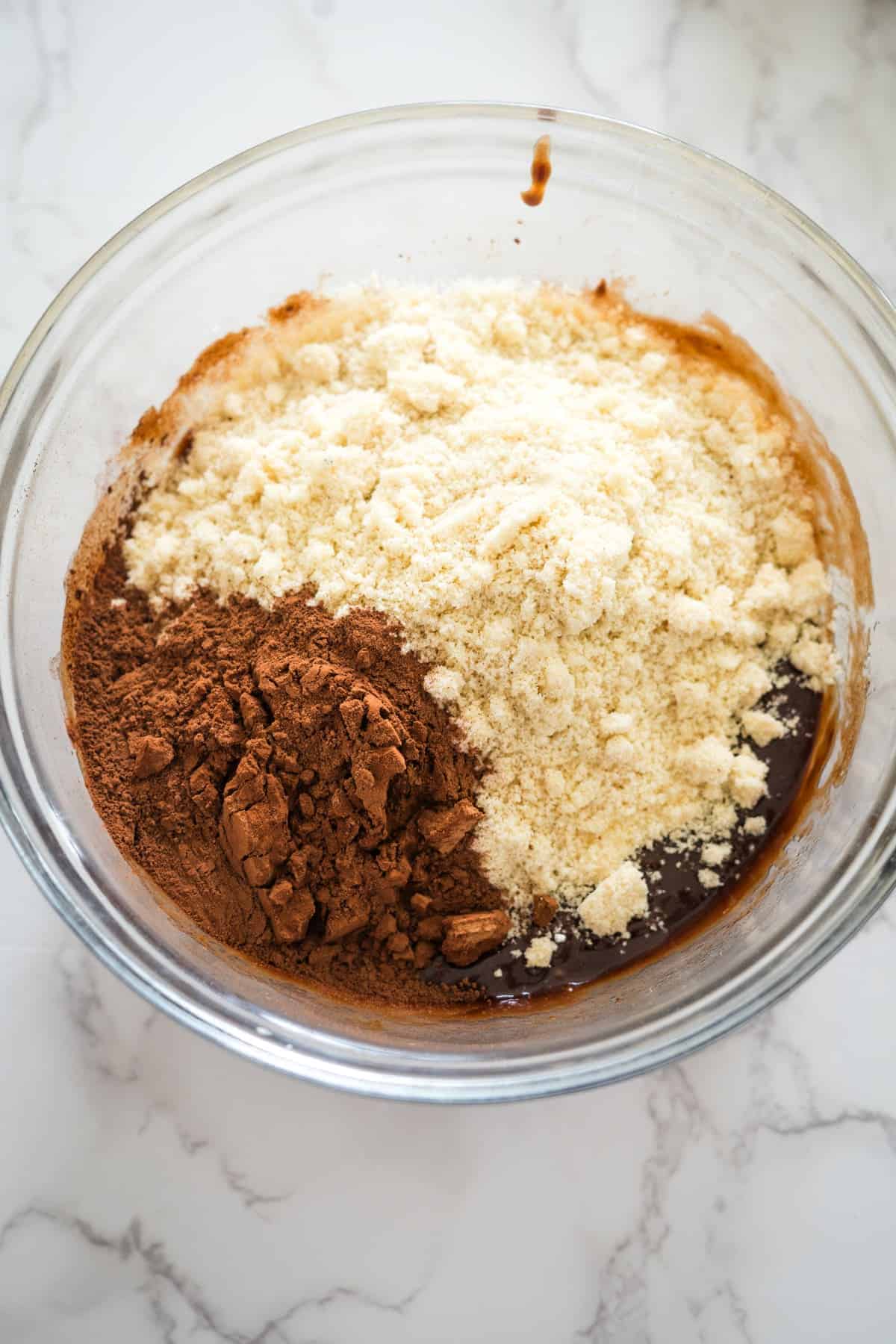 A clear glass bowl containing unsweetened cocoa powder, almond flour, and a liquid mixture, ready to be mixed for a Nutella brownie cheesecake, is placed on a marble countertop.