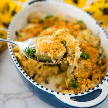 A spoon lifting a serving of creamy baked artichoke dish topped with golden breadcrumbs and garnished with parsley, from a speckled blue dish.