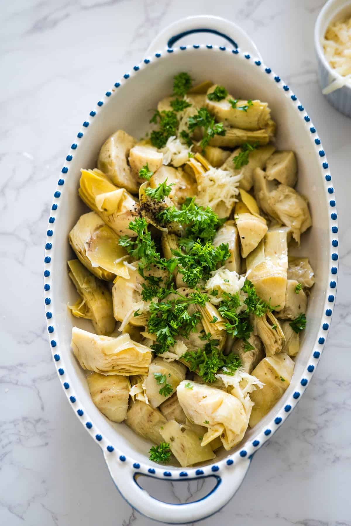 A dish of baked artichoke hearts topped with parsley and grated cheese, served in an oval, blue-dotted ceramic bowl on a marble surface.