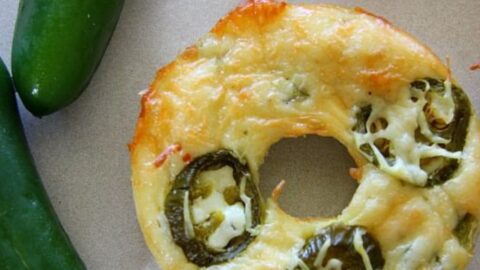 A jalapeno cheese bagel sits on a surface next to three green jalapeno peppers.