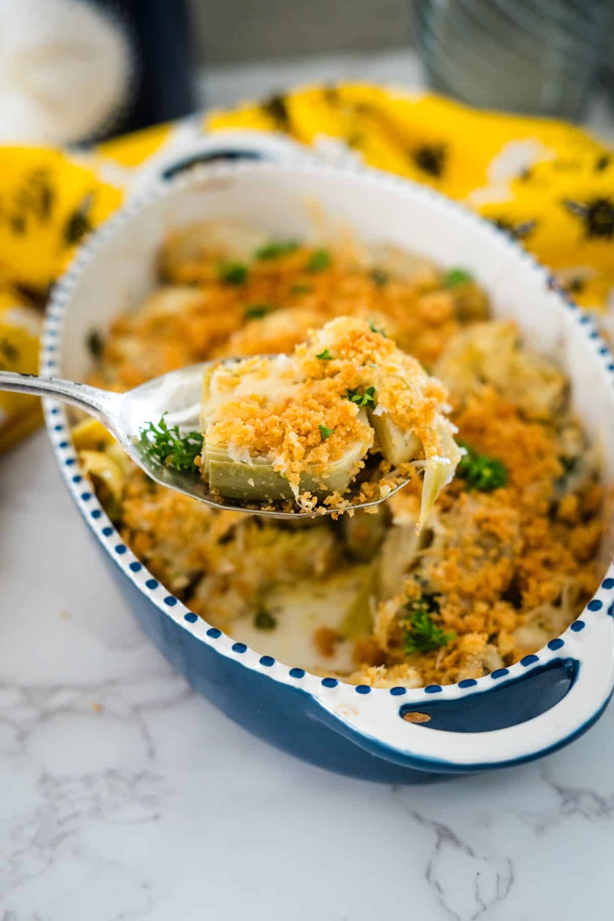 A serving spoon lifts a portion of baked macaroni and cheese with crispy baked artichoke hearts topping from a decorative blue and white dish garnished with parsley.