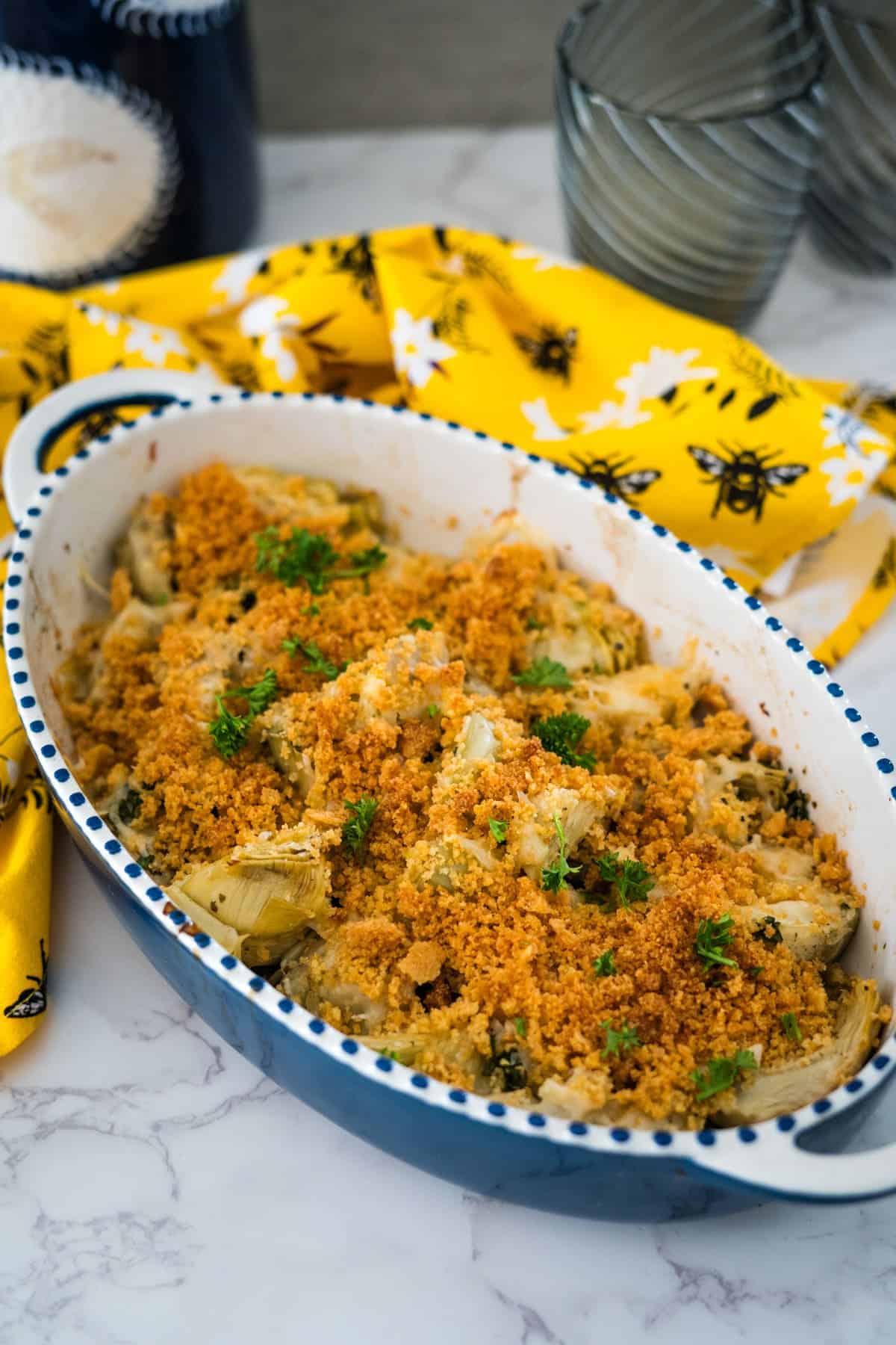 Baked macaroni and cheese in a white and blue speckled dish, garnished with parsley and breadcrumbs, placed on a marble counter with a yellow floral cloth.