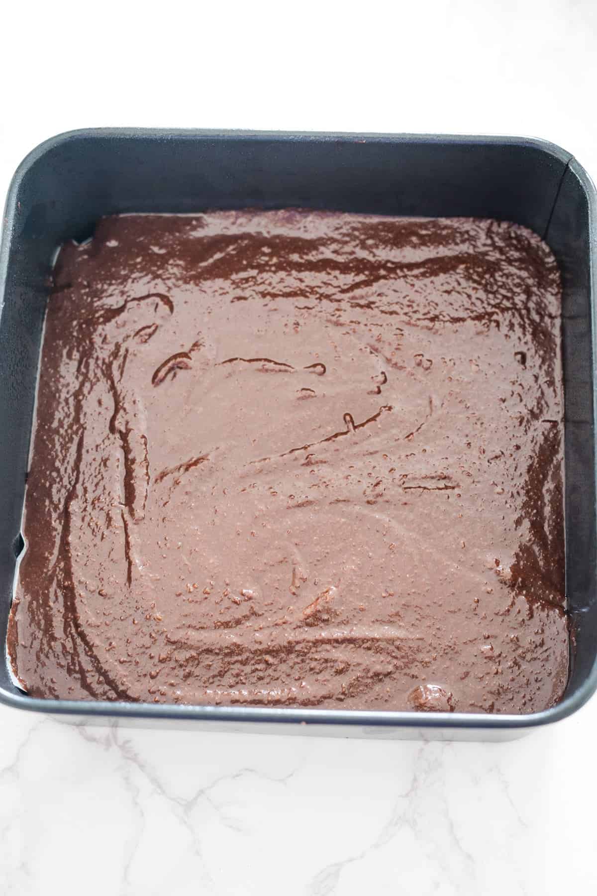 A square baking pan filled with uncooked Nutella brownie batter rests on a white surface, ready to transform into a rich, decadent cheesecake delight.