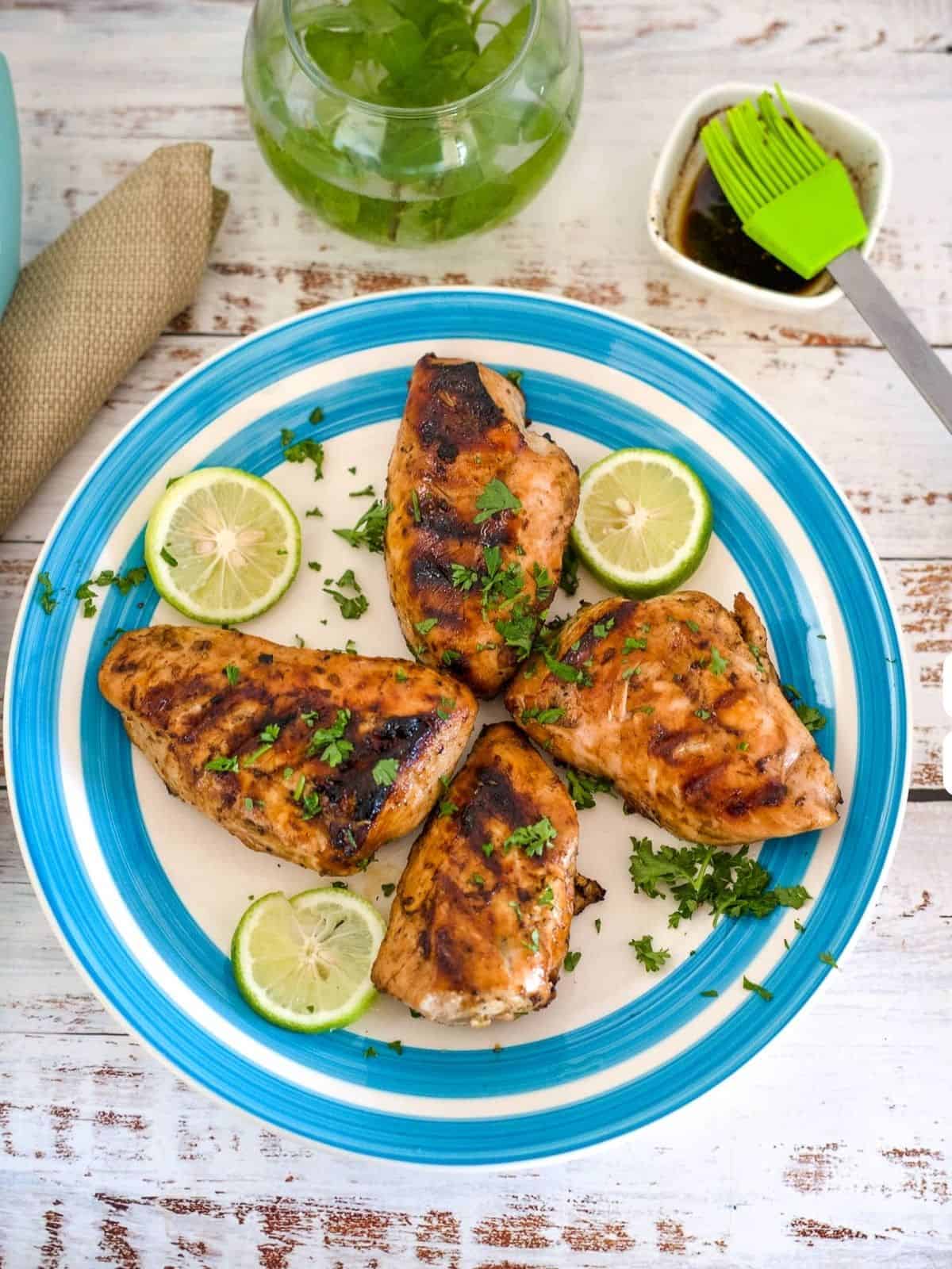 Four grilled chicken breasts garnished with chopped herbs and lime slices are served on a blue and white plate, reminiscent of chicken kofta kebabs. A green basting brush and a bowl of sauce are seen nearby on a wooden table.