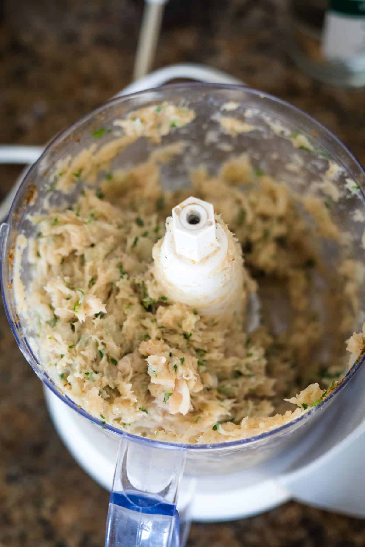 Close-up of a food processor containing a blended mixture with visible herbs on a kitchen countertop, ready to create flavorful chicken kofta kebabs.