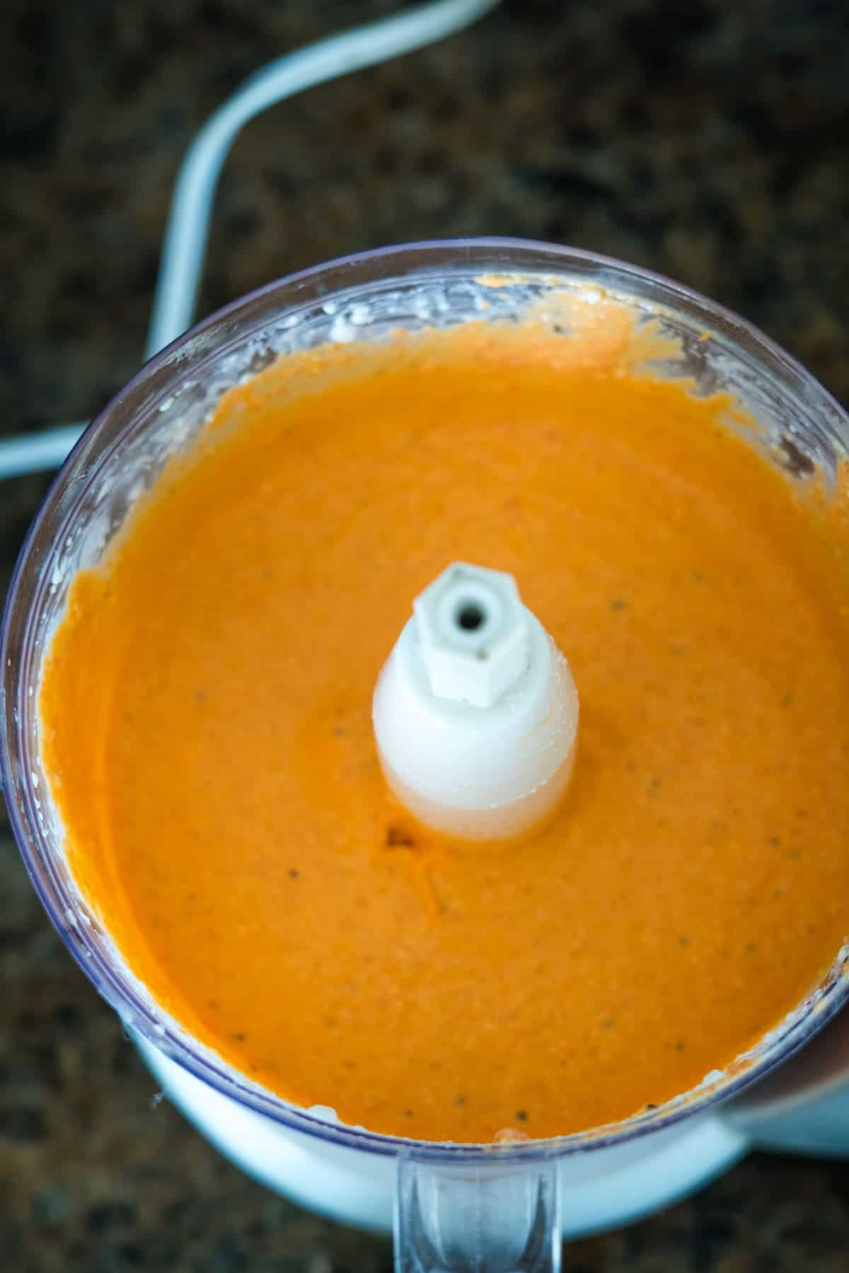 Top view of a food processor containing blended orange sauce, set on a dark countertop.