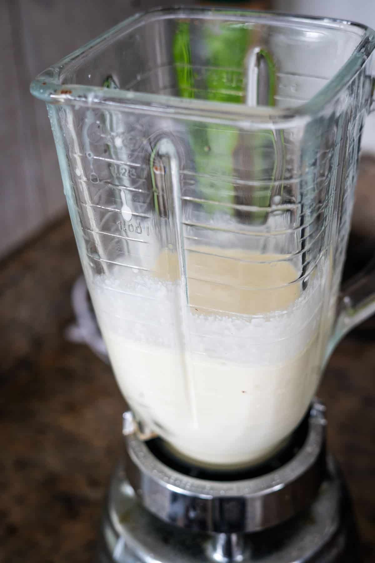 A glass blender jar containing a mixture of white liquid with a layer of white powder on top, sitting on a metallic blender base on a brown countertop.