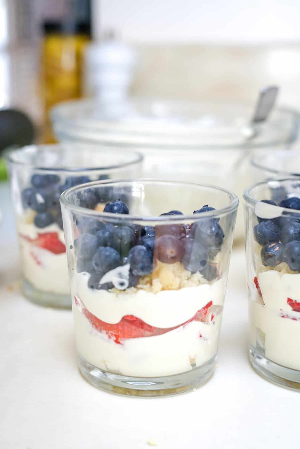 Three glass cups filled with layers of cream, sliced strawberries, crumbled cookies, and fresh blueberries on top sit on a countertop. A spoon is visible in a bowl in the background.