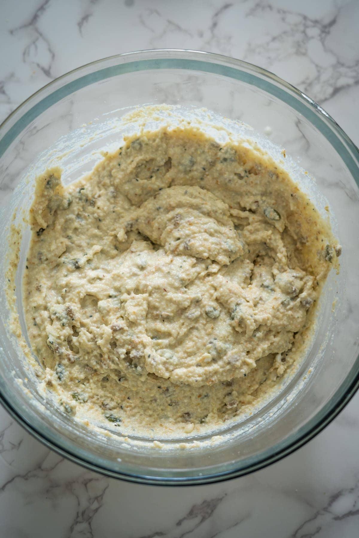 A glass bowl filled with a creamy mixture containing what appears to be ricotta cheese with herbs on a marble countertop.