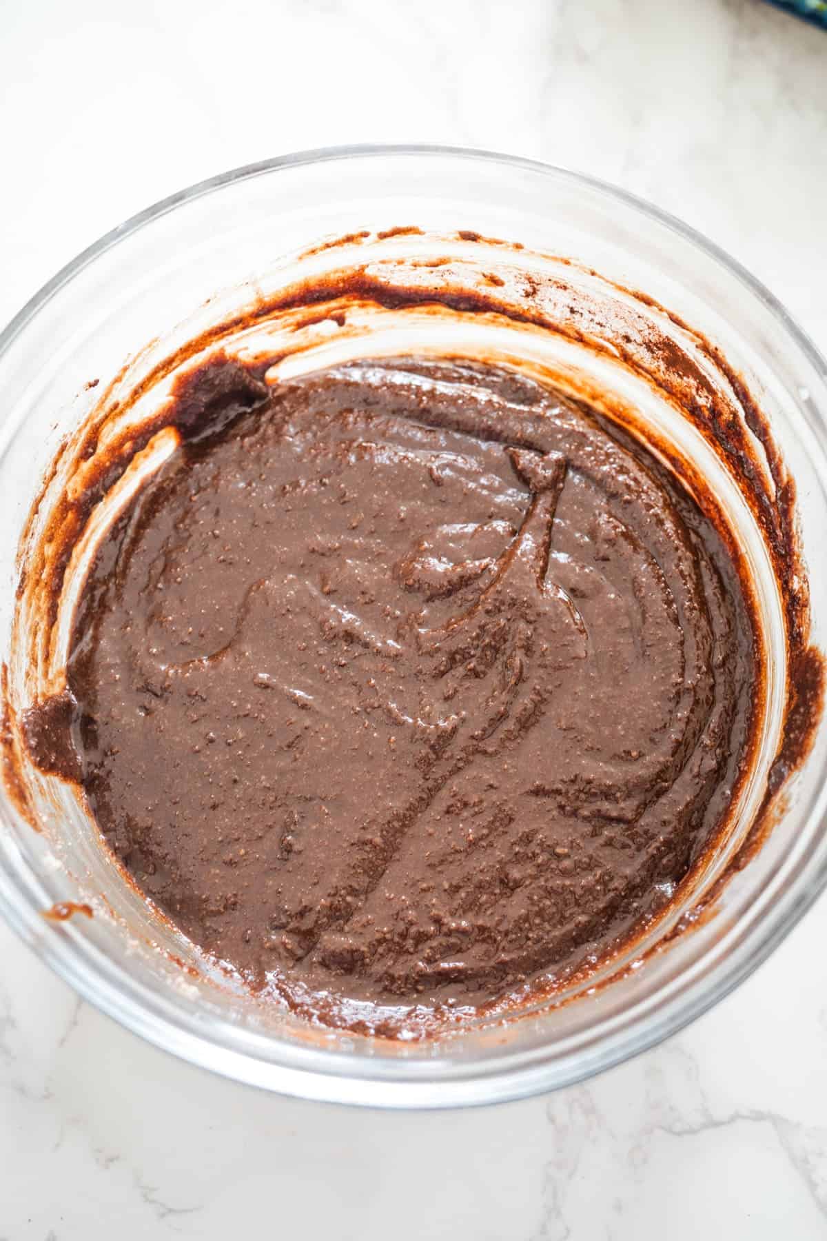 A glass bowl filled with rich, chocolate batter for a Nutella brownie cheesecake rests on a white marble countertop.