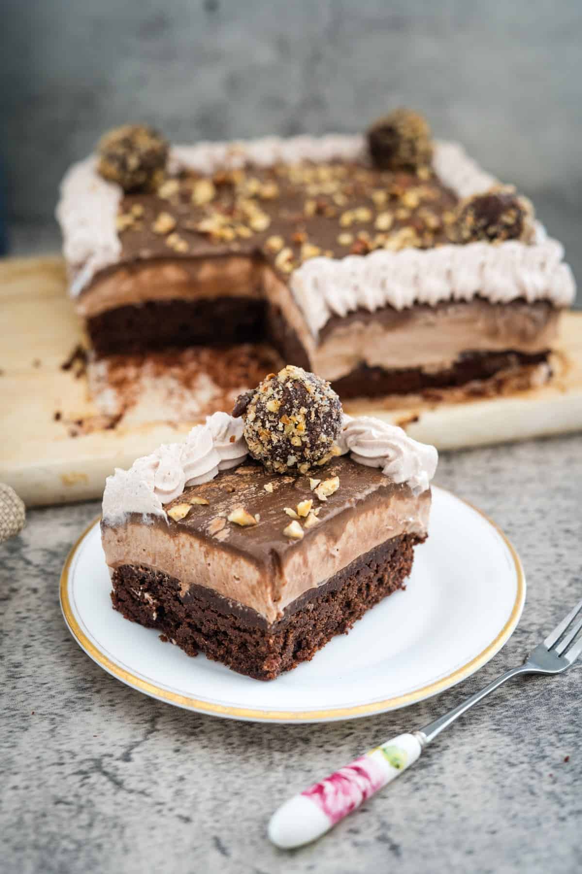 A piece of layered chocolate cake with whipped cream topping and nut garnish on a white plate, with the remaining Nutella brownie cheesecake in the background on a cutting board.