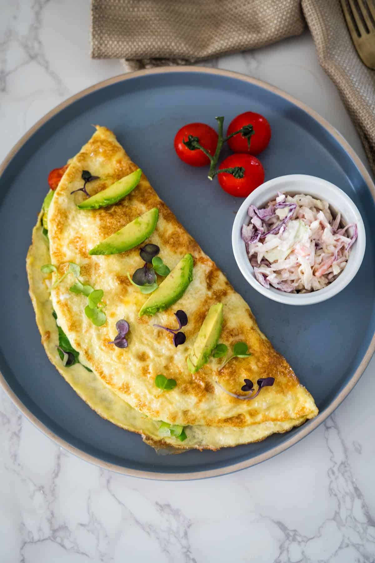 A folded California omelette topped with avocado slices and herbs, served with cherry tomatoes and coleslaw on a blue plate.