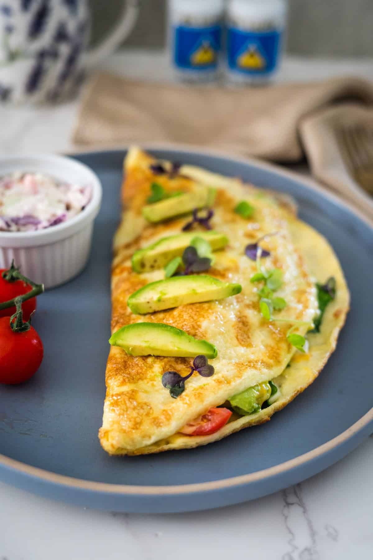 A plate with an omelette topped with avocado slices and microgreens, served with a side of coleslaw and cherry tomatoes.