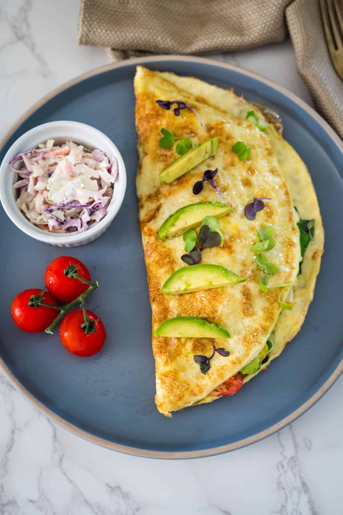 Omelette with avocado slices on a blue plate, accompanied by cherry tomatoes and a small bowl of coleslaw.