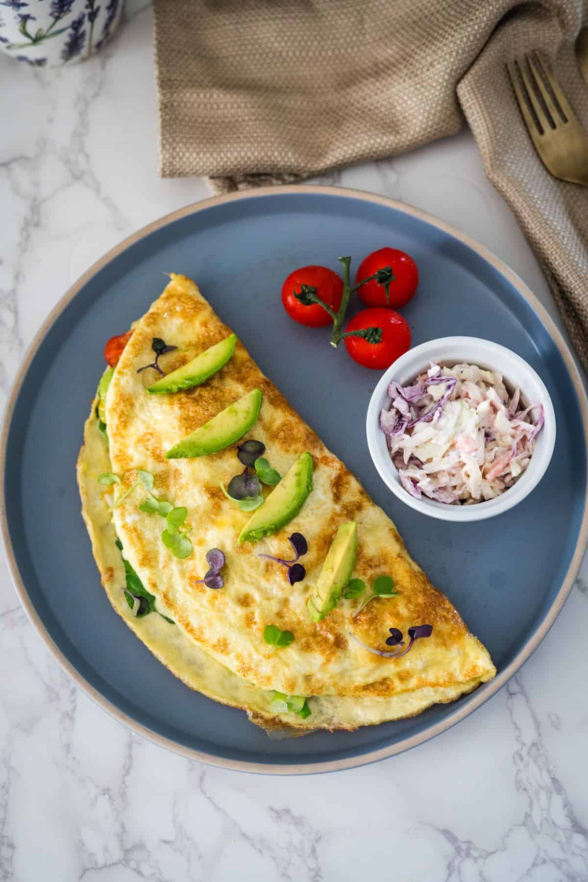 An omelette garnished with avocado and herbs on a blue plate, accompanied by a side of coleslaw and cherry tomatoes.