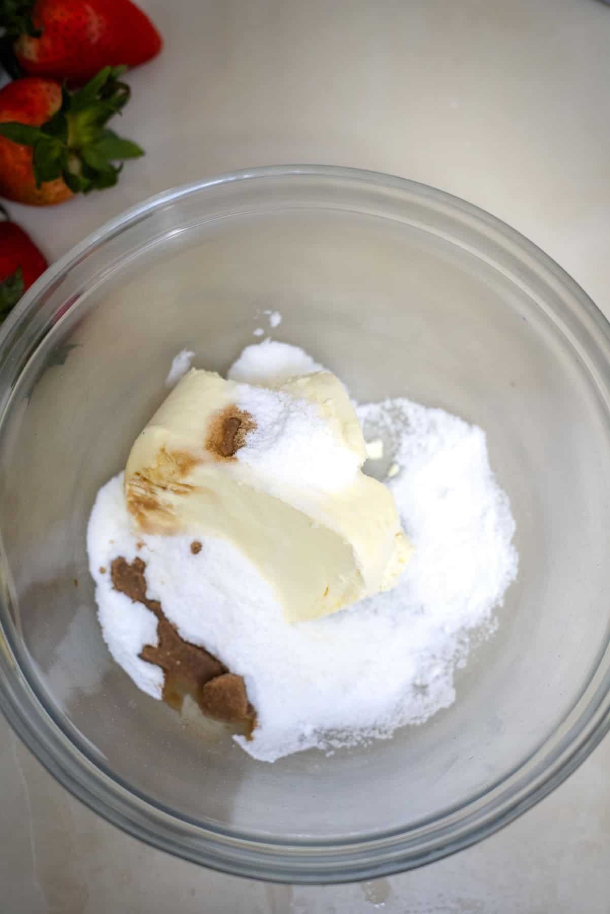 A glass bowl containing butter, white sugar, and brown sugar on a light-colored countertop. Fresh strawberries are visible in the background.