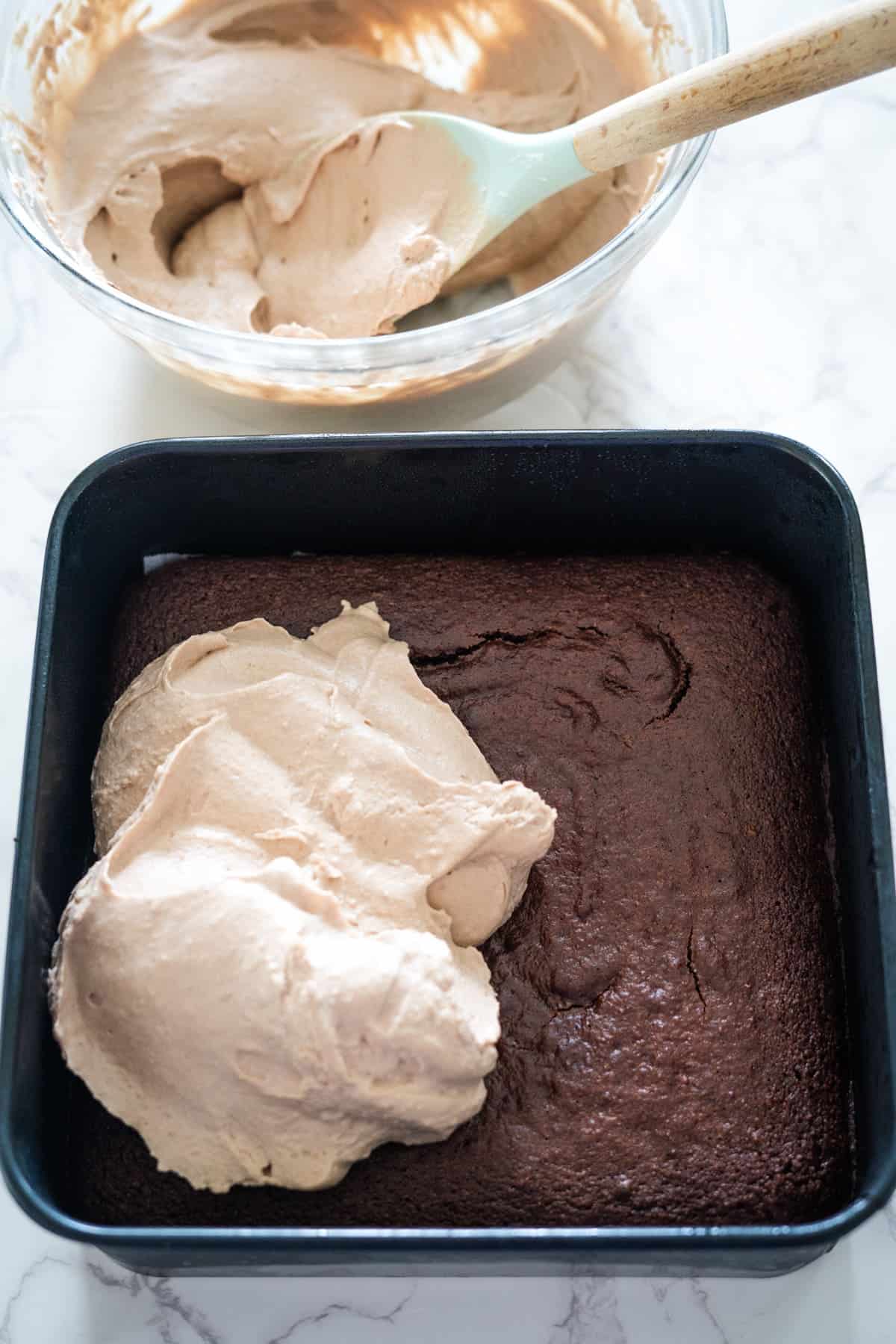 A square Nutella brownie cheesecake in a baking pan with a dollop of light brown frosting on top. A bowl with more frosting and a spatula is visible in the background.