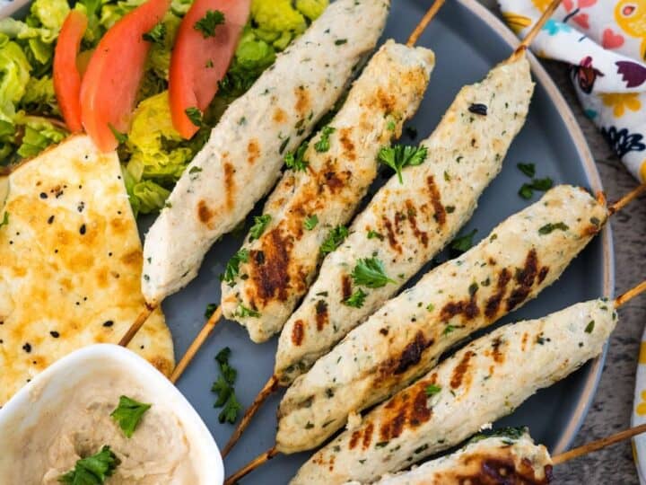 A plate of grilled chicken kebabs on skewers, served with a side salad, slices of tomato, flatbread, and a small dish of dipping sauce.