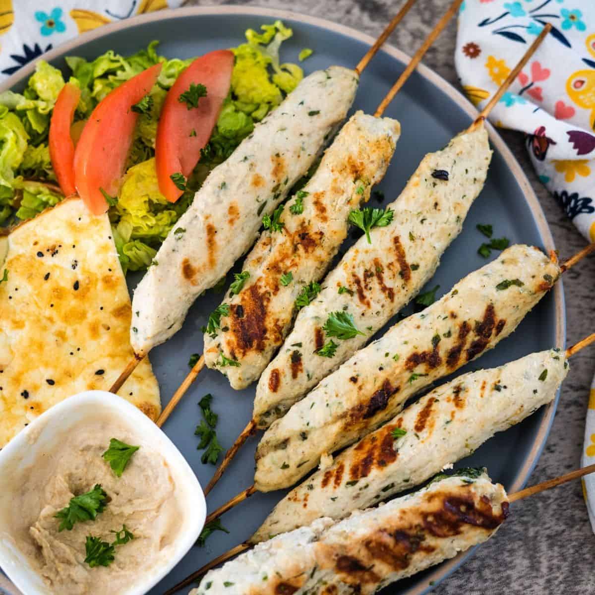 A plate of grilled chicken kebabs on skewers, served with a side salad, slices of tomato, flatbread, and a small dish of dipping sauce.