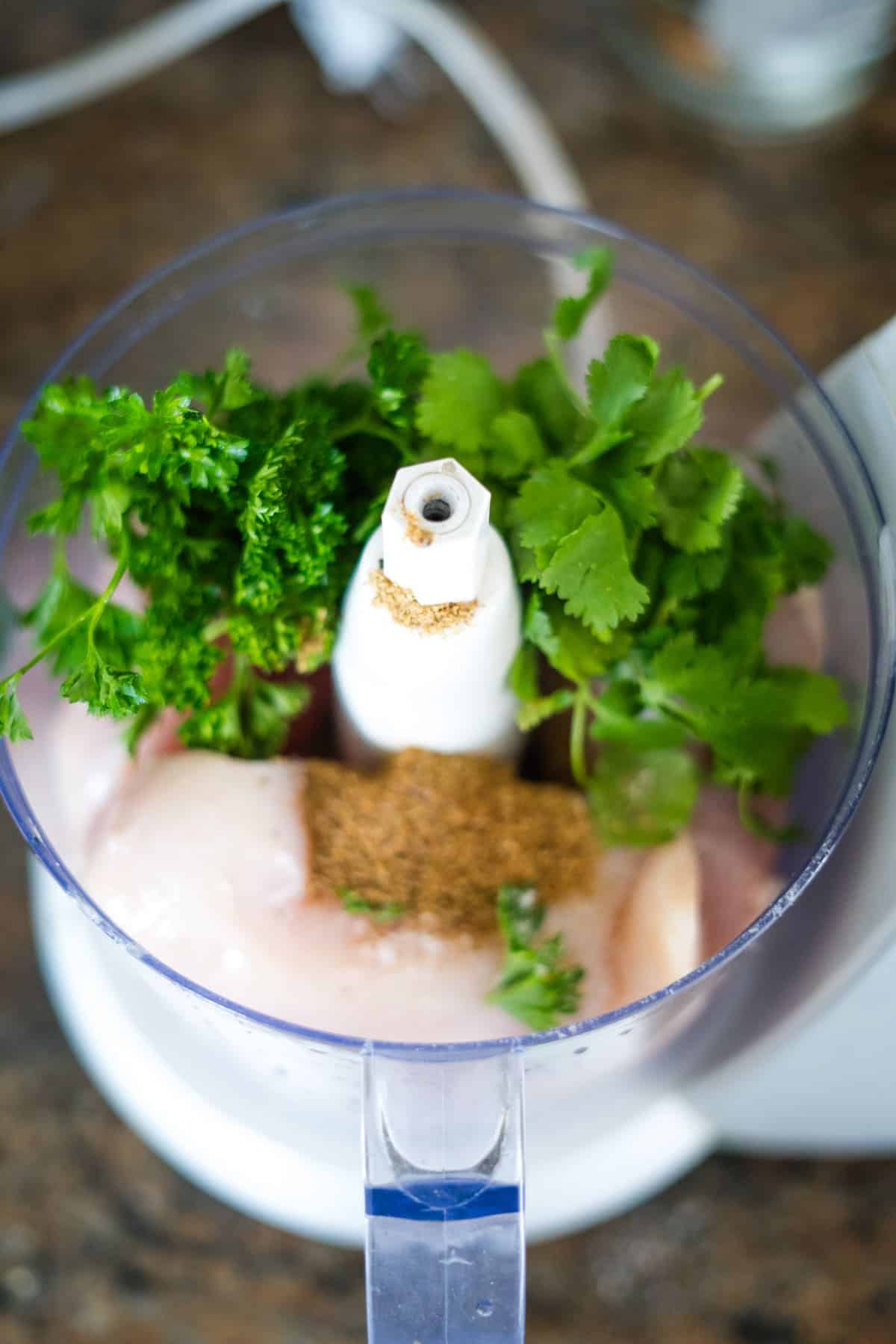 Close-up of a food processor containing raw chicken, fresh parsley, cilantro, and spices for chicken kofta kebabs, viewed from above.