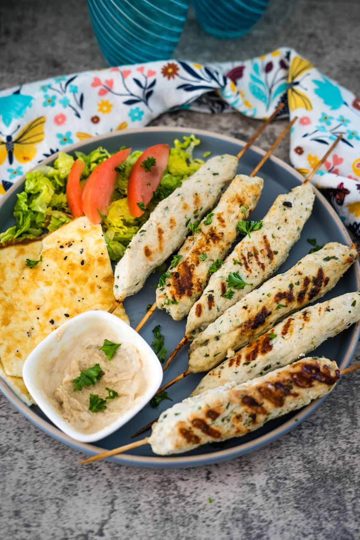 A plate with grilled chicken skewers, a fresh salad with tomato slices, a piece of flatbread, and a small dish of dip, garnished with fresh herbs and sesame seeds.