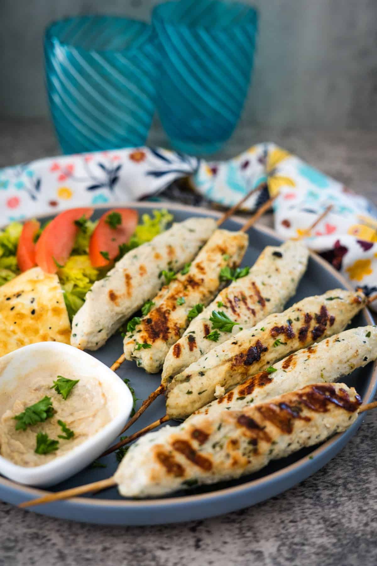 A plate with chicken kofta kebabs, hummus, sliced tomatoes, yellow rice, and pita bread, alongside two blue glasses and a patterned napkin.