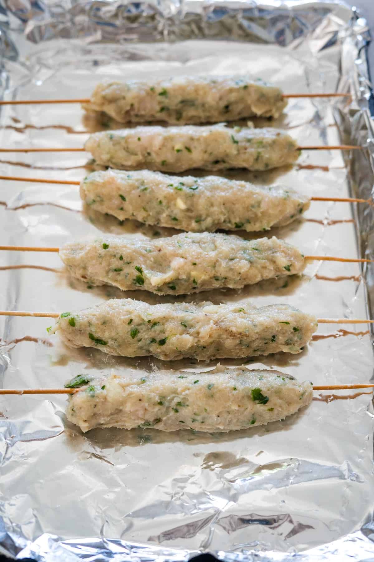 Six uncooked chicken kofta kebabs, made from a mixture of chicken and herbs, are laid out on skewers on a foil-lined baking sheet.