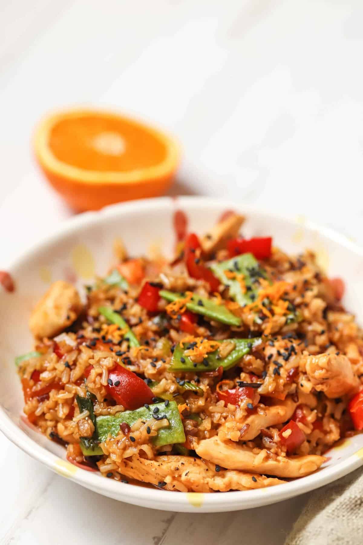 A bowl of stir-fried rice with chicken, red bell peppers, green beans, and sprinkled sesame seeds, with a halved orange in the background.