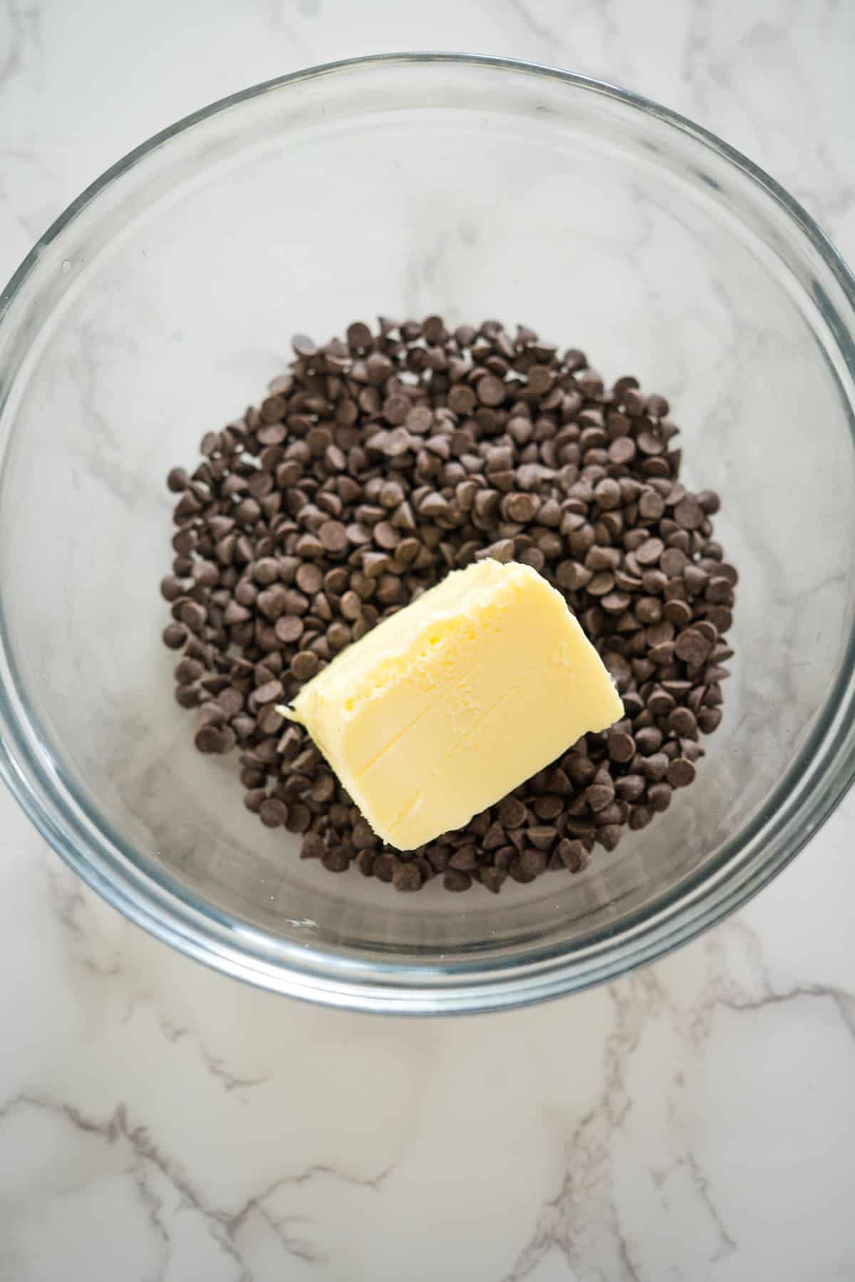 A glass bowl contains chocolate chips and a block of butter on a marble surface, setting the stage for a decadent Nutella brownie cheesecake.