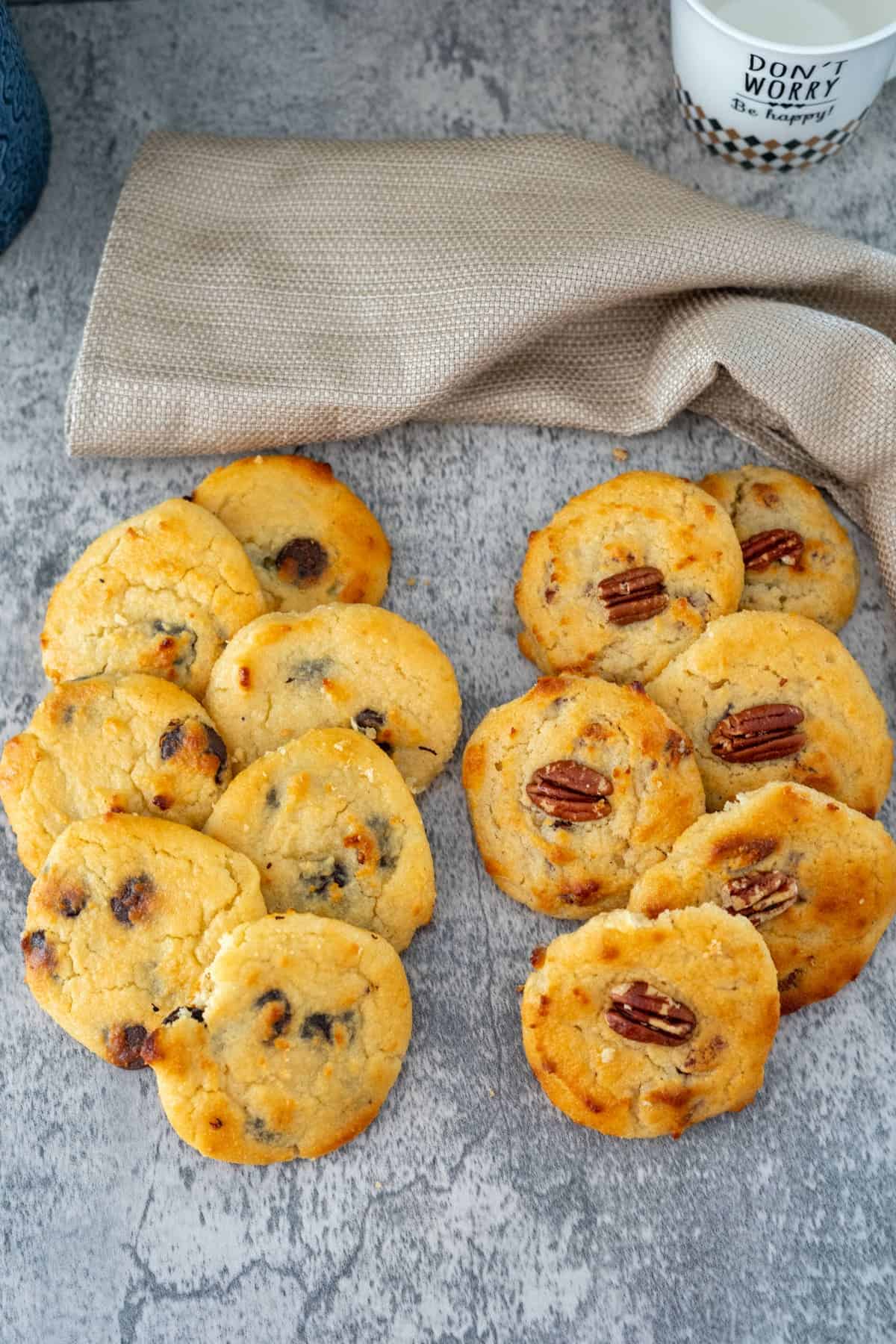 A group of assorted cookies, including delightful cottage cheese cookies, some with chocolate chips and others topped with pecans, arranged on a gray surface near a beige cloth and a portion of a white mug.