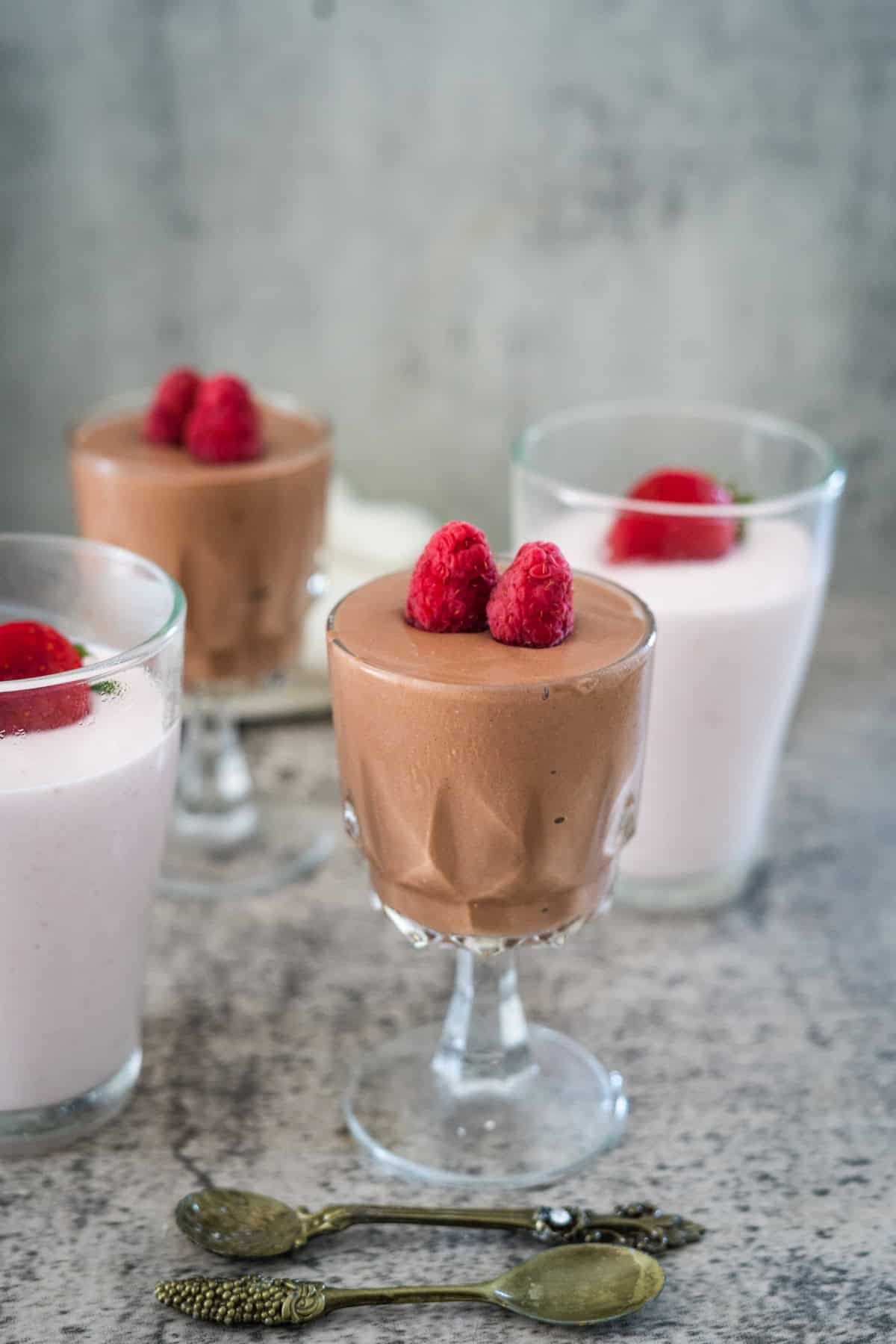 Three glasses of mousse, two chocolate and one strawberry, each topped with raspberries. Two decorative spoons are in the foreground.