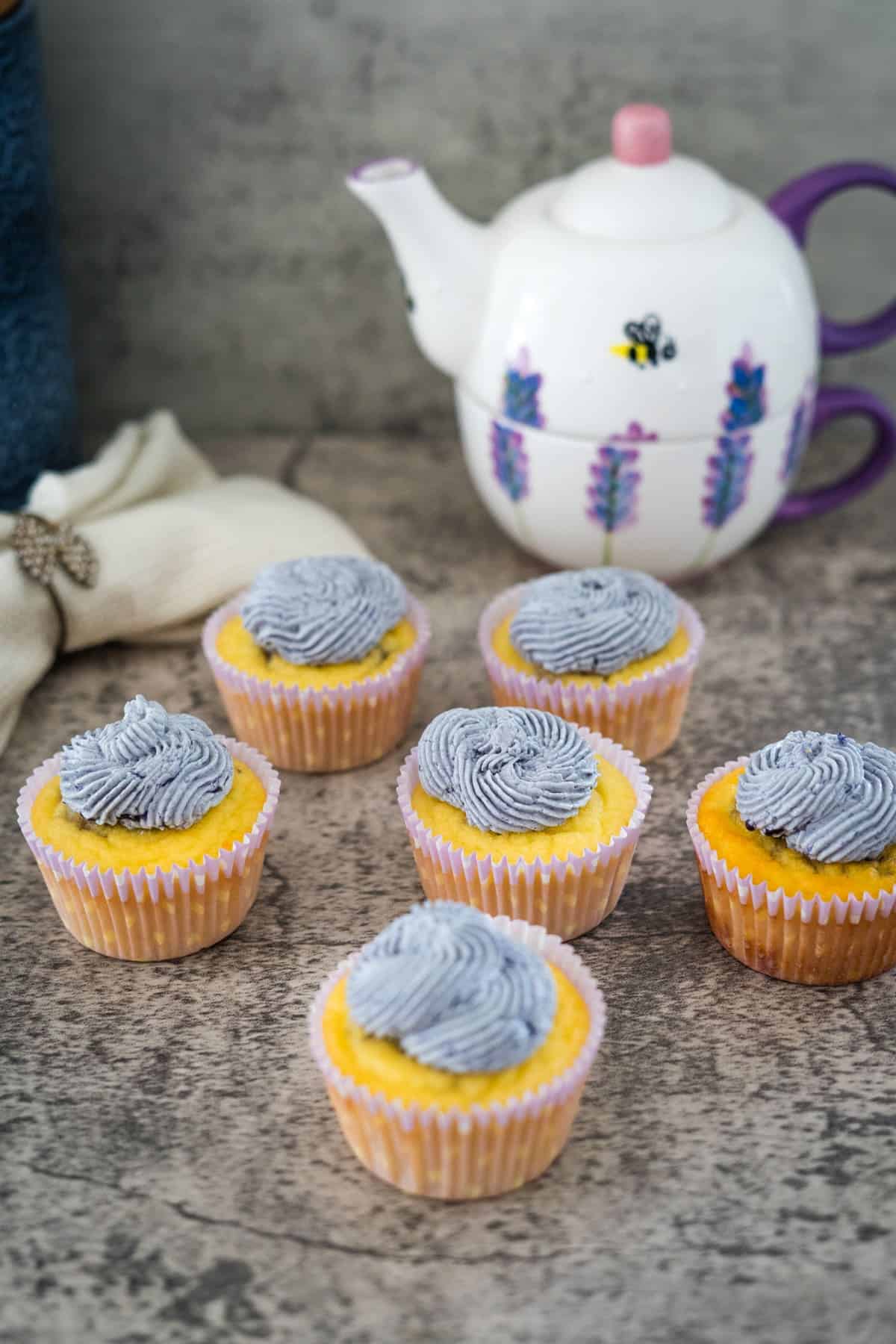 Six lemon lavender cupcakes with blue frosting are arranged on a stone-like surface. A white teapot with lavender and bee designs is in the background.