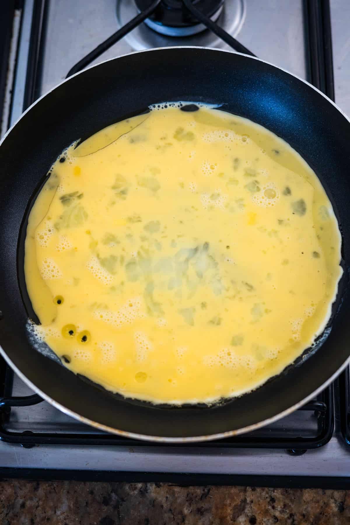 A mixture of beaten eggs and chopped herbs cooking in a black non-stick skillet on a stove.