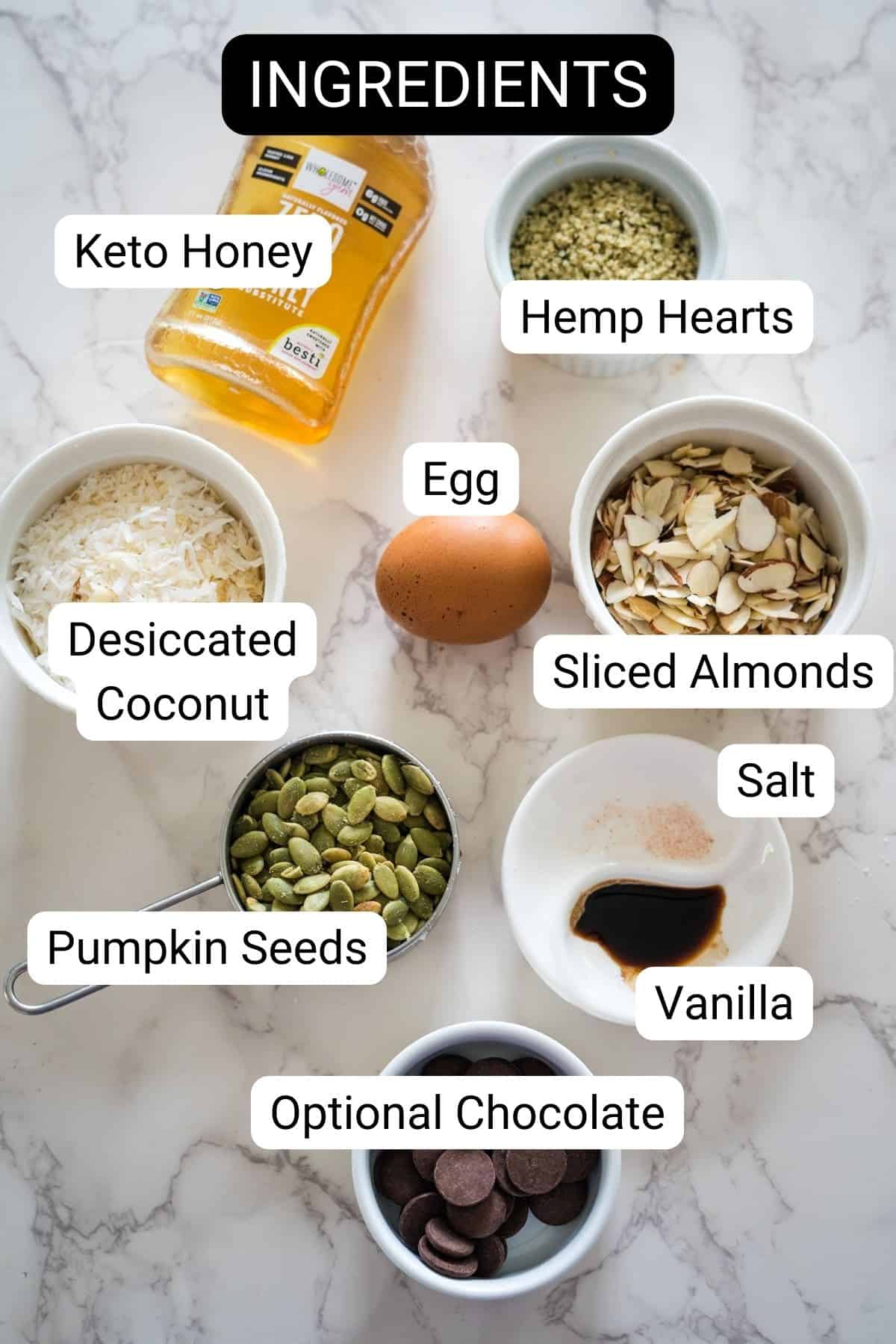 Assorted baking ingredients labeled on a marble surface for keto breakfast cookies, including keto honey, hemp hearts, egg, desiccated coconut, sliced almonds, pumpkin seeds, salt, vanilla, and optional chocolate.