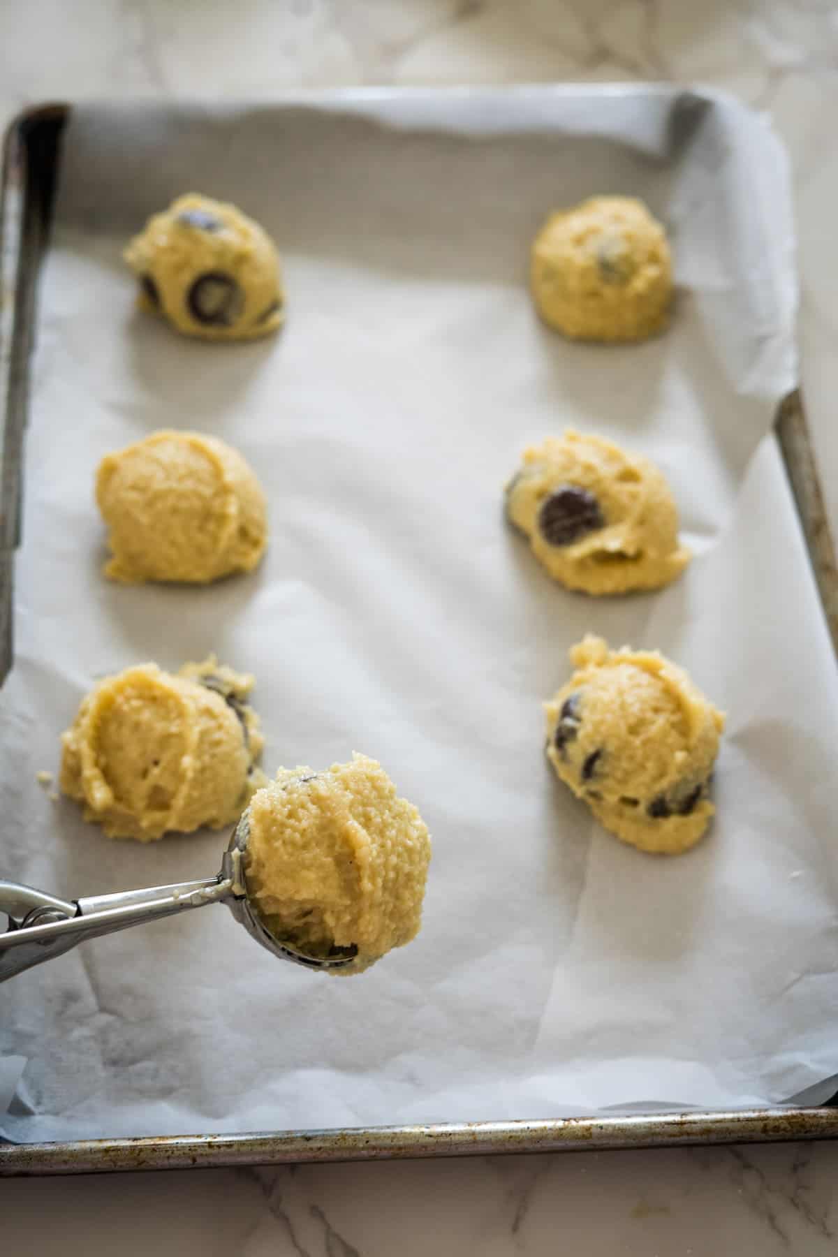 A baking tray lined with parchment paper holds six mounds of cottage cheese cookie dough. An ice cream scoop with one additional dough mound is shown in the foreground.