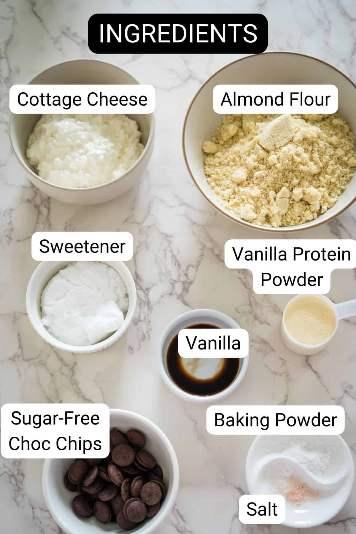 A collection of labeled ingredients including cottage cheese, almond flour, sweetener, vanilla protein powder, vanilla, baking powder, sugar-free chocolate chips, and salt on a marble surface.