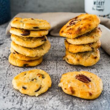 Two stacks of homemade cookies with mixed nuts and chocolate chips are placed on a rustic grey surface, with a few cookies lying beside the stacks. A beige cloth and a bottle are in the background.