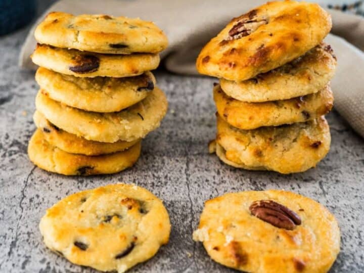 Two stacks of homemade cookies with mixed nuts and chocolate chips are placed on a rustic grey surface, with a few cookies lying beside the stacks. A beige cloth and a bottle are in the background.