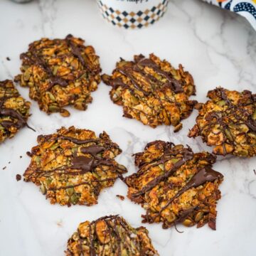 Homemade granola cookies with seeds and nuts, drizzled with dark chocolate, on a marble surface.