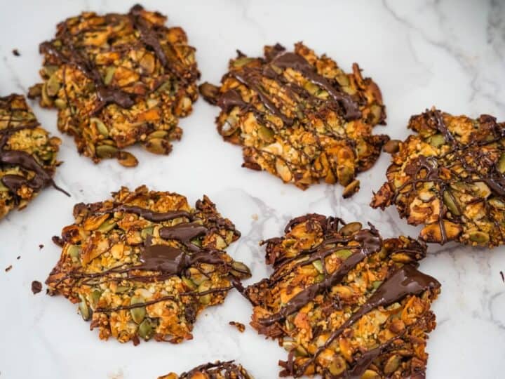 Homemade granola cookies with seeds and nuts, drizzled with dark chocolate, on a marble surface.