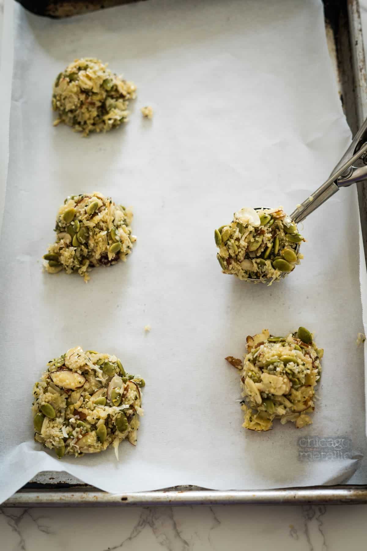 Scoops of keto cookie dough with seeds and oats on a baking sheet lined with parchment paper, using a metal scoop.