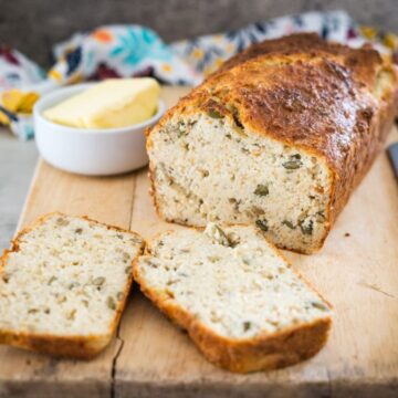 A loaf of zucchini bread with two slices cut, displayed on a wooden board. A small bowl of butter is placed next to the loaf.