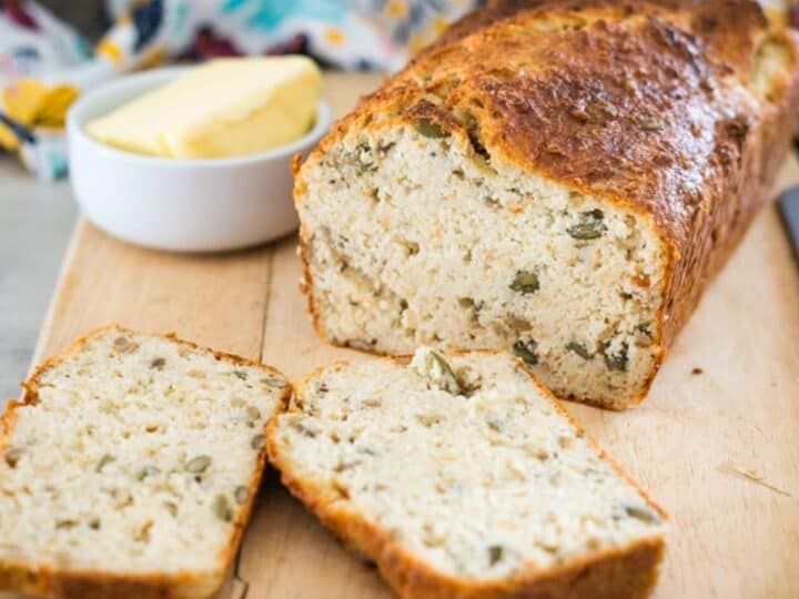 A loaf of zucchini bread with two slices cut, displayed on a wooden board. A small bowl of butter is placed next to the loaf.