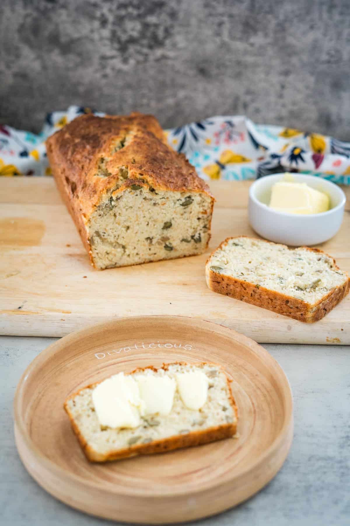 A loaf of sliced cottage cheese bread sits on a wooden cutting board with a colorful cloth in the background. Nearby, a plate holds a partially buttered slice, with a small bowl of butter completing the scene.