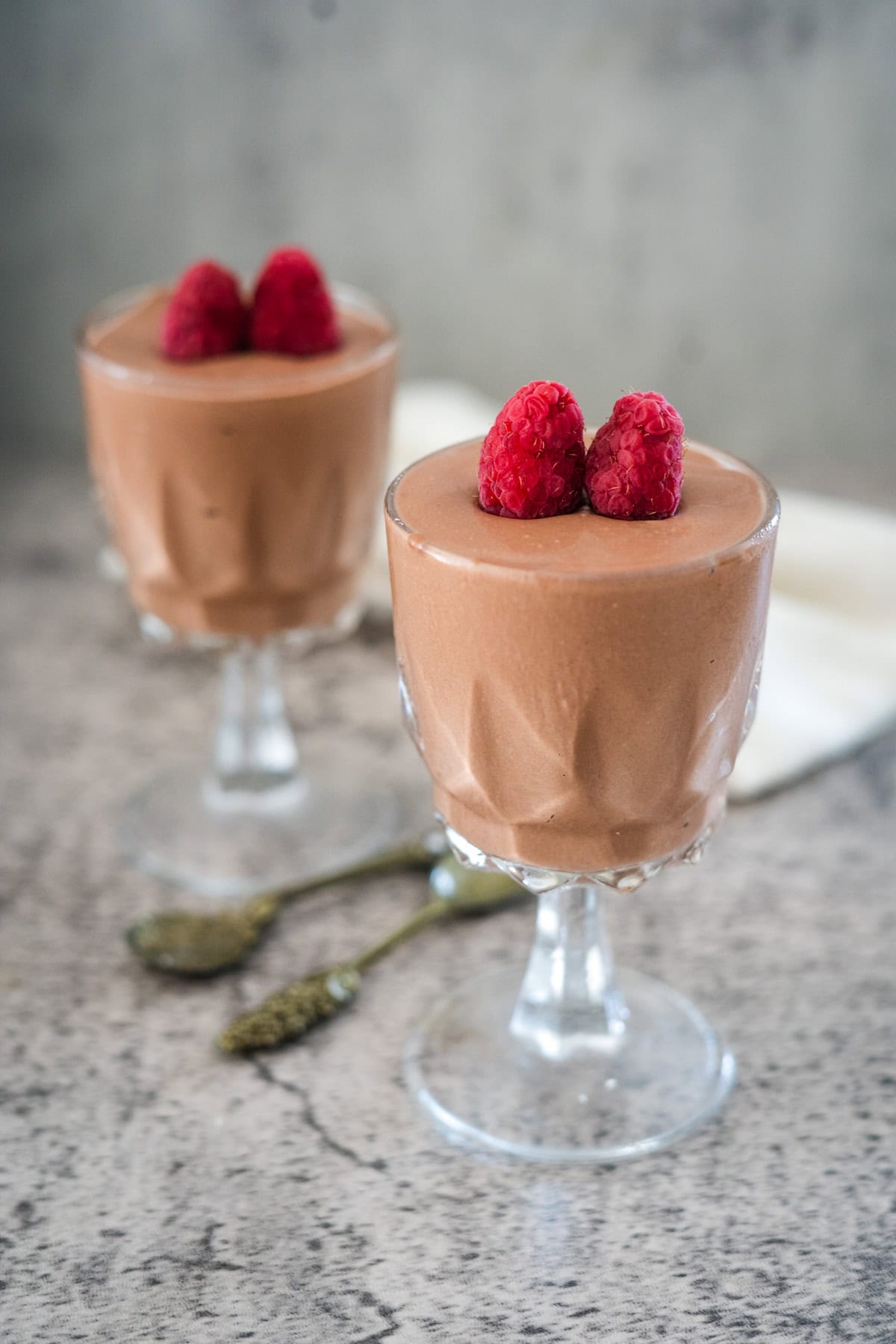 Two glass cups of chocolate mousse topped with raspberries, placed on a textured surface with two small spoons between them.