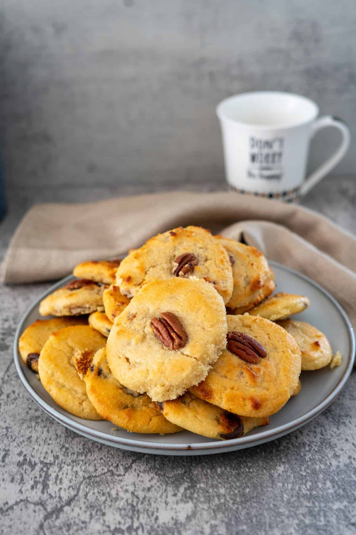 A plate of homemade cookies topped with nuts is placed on a gray surface with a beige napkin and a white coffee mug in the background.