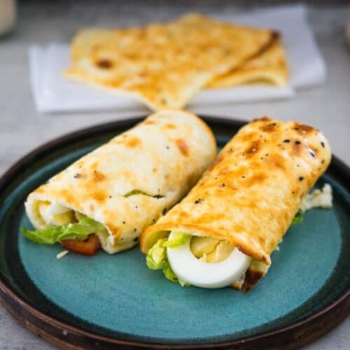 Two breakfast wraps containing boiled eggs, lettuce, and other ingredients are served on a teal plate with more wraps in the background.