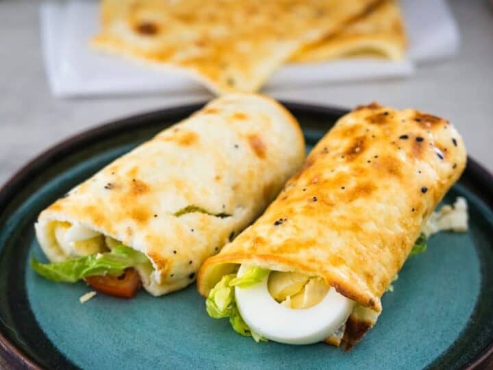 Two breakfast wraps containing boiled eggs, lettuce, and other ingredients are served on a teal plate with more wraps in the background.